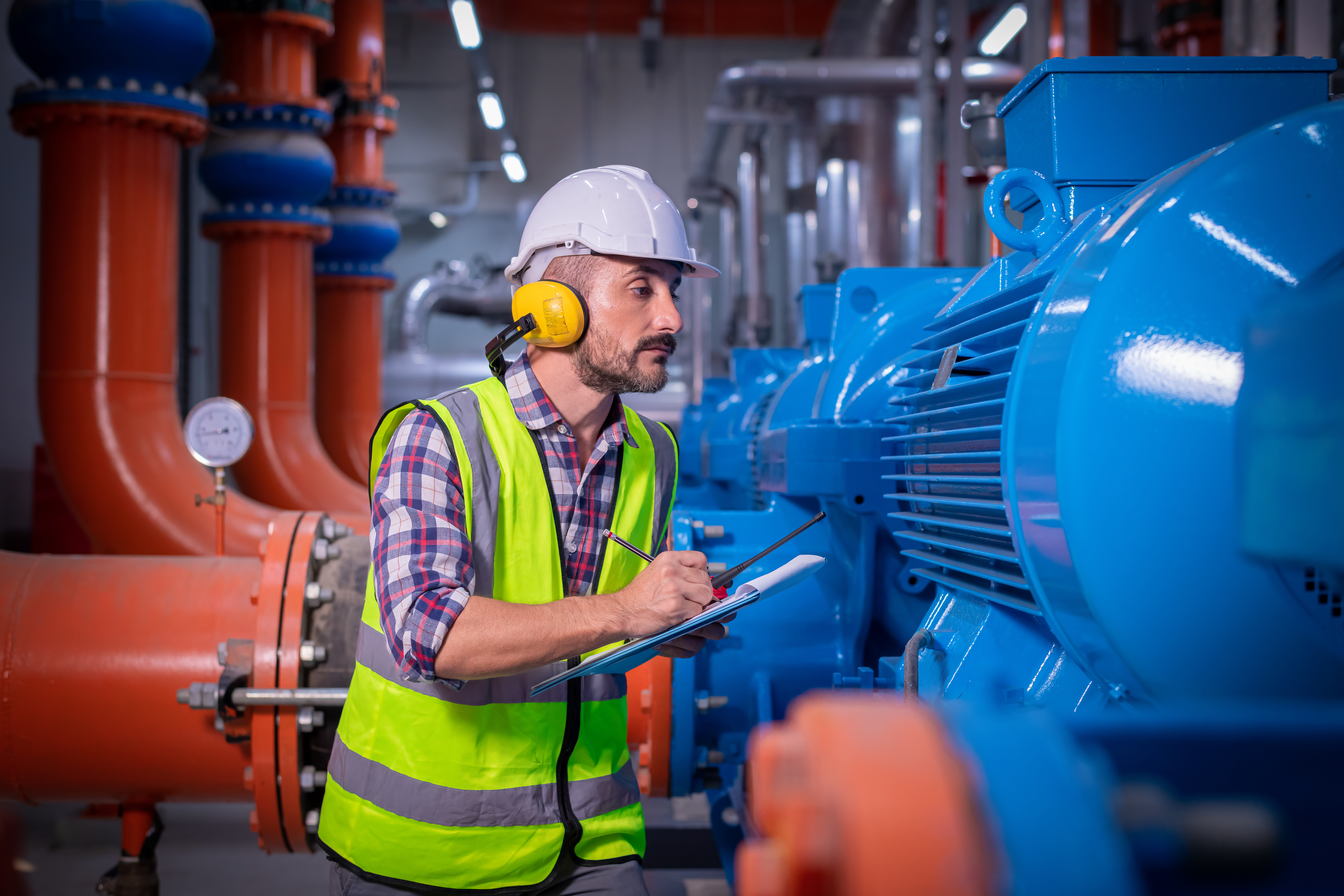 A technician wearing a white hard hat, yellow safety vest, and ear protection inspects a large industrial air chiller. He is holding a clipboard and pen, surrounded by vibrant blue machinery and orange piping, representing HVAC maintenance and system efficiency checks.