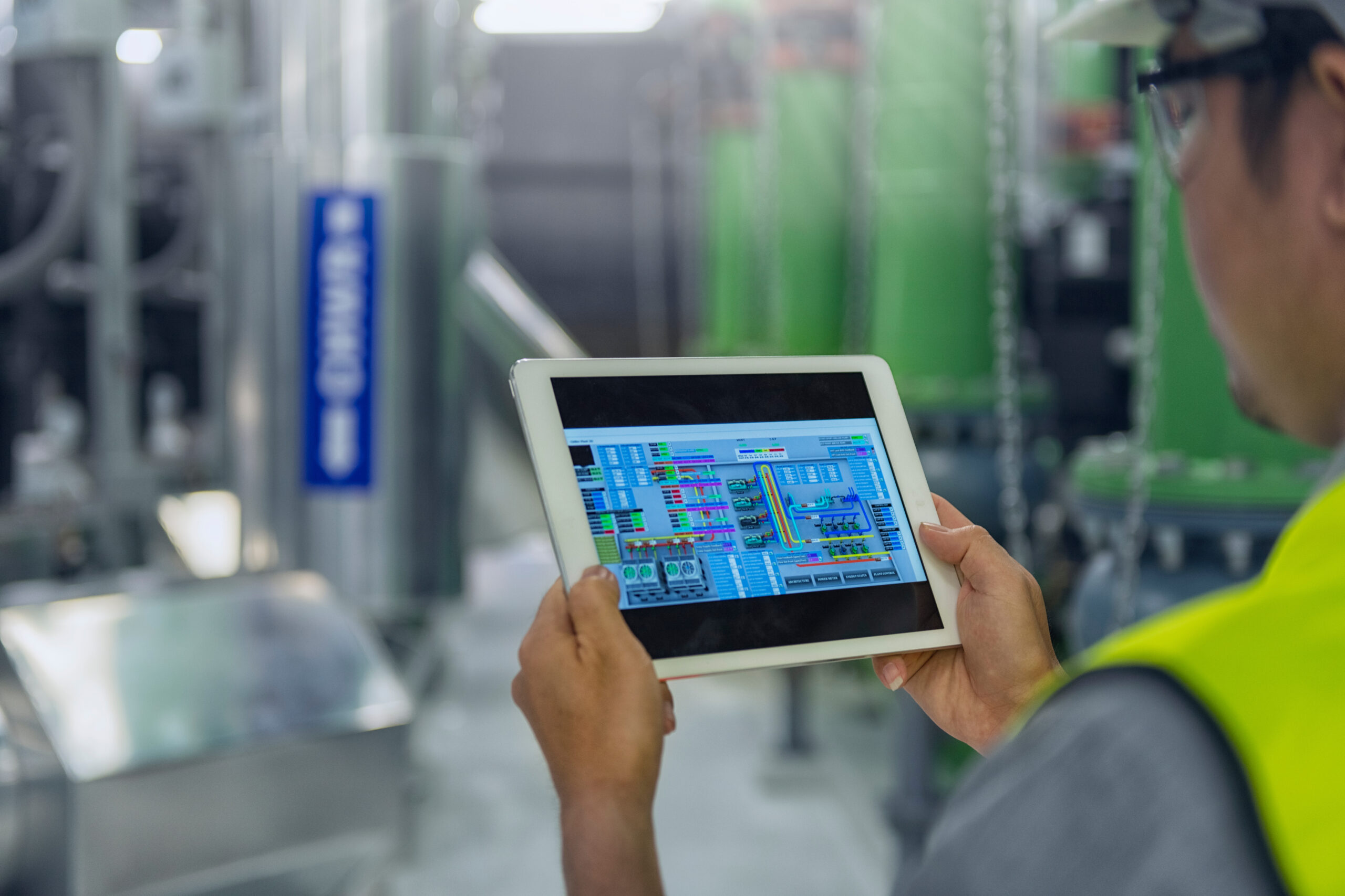 A technician wearing a high-visibility vest and safety glasses operates a tablet displaying a detailed building automation system interface, surrounded by industrial equipment, symbolizing smart technology integration and efficient facility management