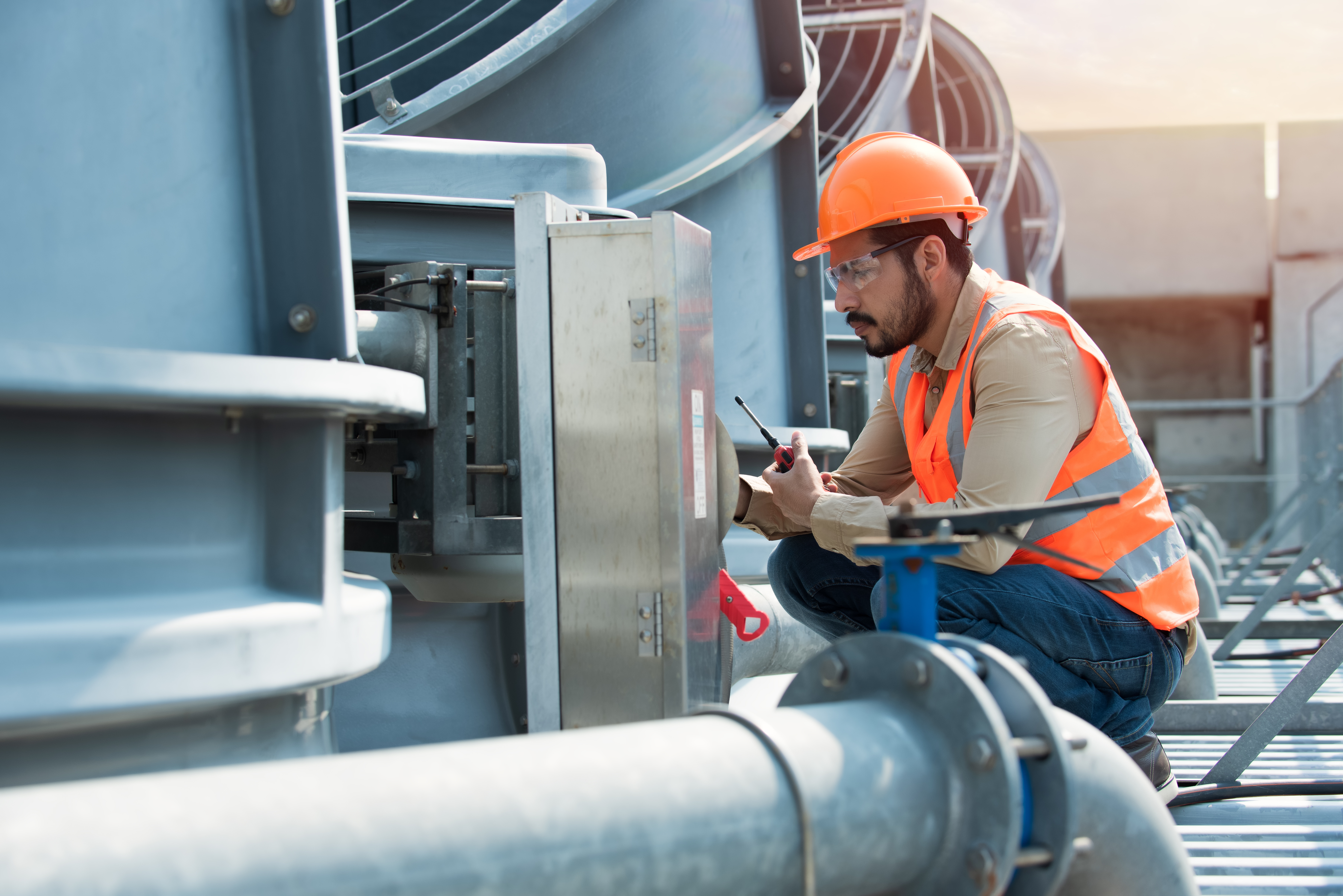 A male engineer wearing an orange safety helmet, reflective vest, and safety glasses inspects industrial HVAC equipment outdoors. He is kneeling beside large metal pipes, using tools to perform maintenance or repairs, with sunlight softly illuminating the scene.