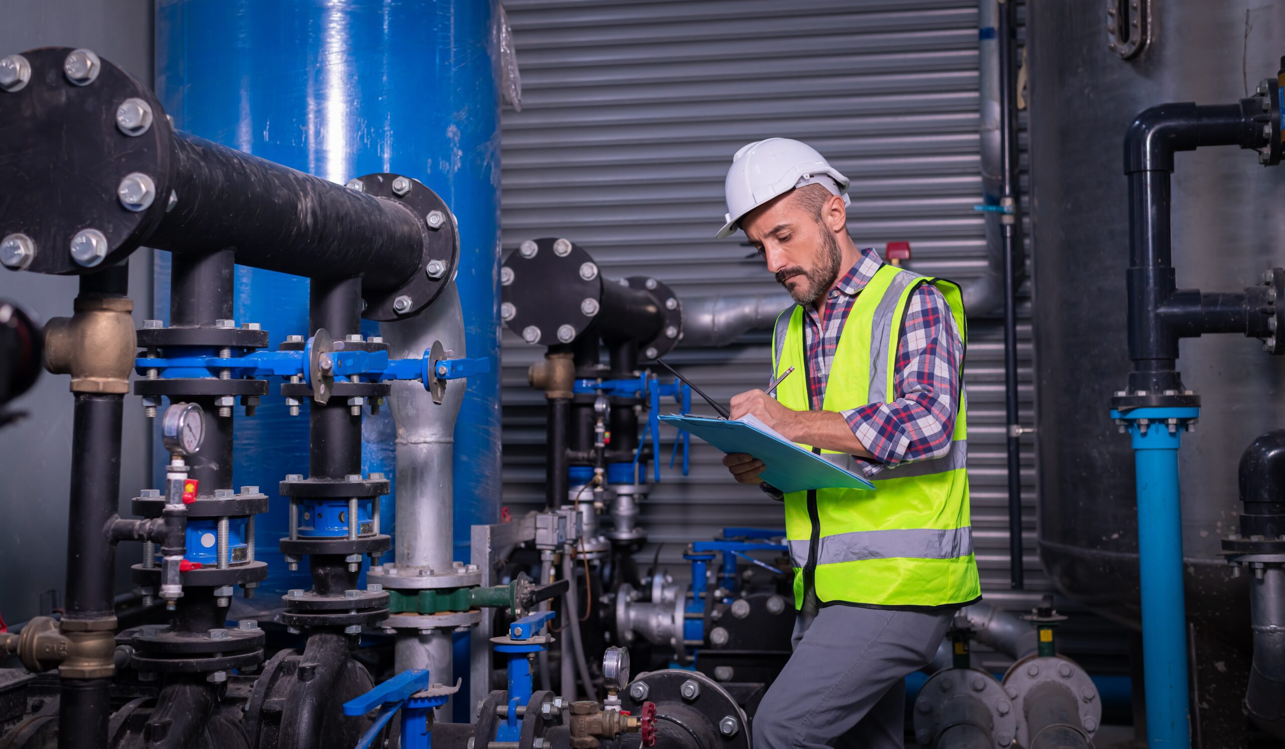 A male technician in a white hard hat and reflective vest inspects industrial piping, making notes on a clipboard in a mechanical room filled with large pipes and valves.